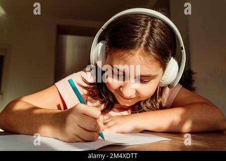 Lächelndes Mädchen mit kabellosen Kopfhörern, das am Schreibtisch ein Buch schreibt Stockfoto
