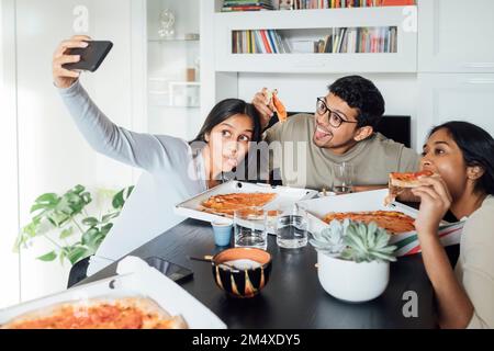 Junge Frau macht Selfie über Handy, während Frau und Mann zu Hause Pizza essen Stockfoto