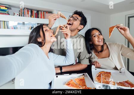 Eine glückliche junge Frau macht Selfie mit einer Frau und einem Mann, die zu Hause Pizza essen Stockfoto