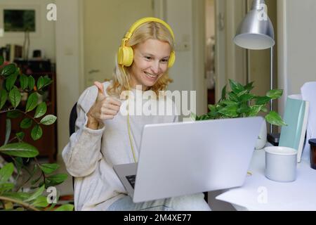 Lächelnde Frau mit Kopfhörern, die zu Hause auf dem Laptop sitzt Stockfoto