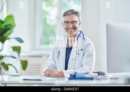 Glücklicher reifer Arzt im Labormantel sitzt in der Praxis Stockfoto