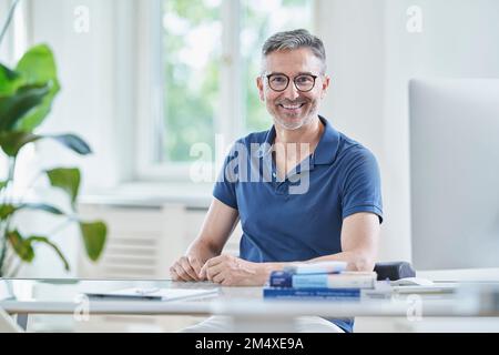 Glücklicher Arzt mit Brille sitzt am Schreibtisch in der Praxis Stockfoto