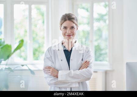 Lächelnde Ärztin im Labormantel mit gekreuzten Armen in der Praxis Stockfoto