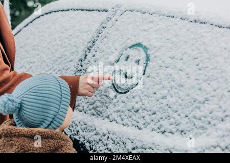 Ein Junge, der neben der Mutter steht und ein menschliches Gesicht auf einem schneebedeckten Autofenster malt Stockfoto