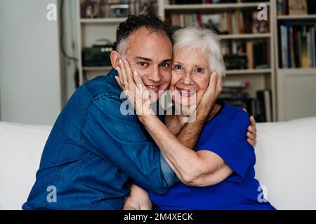 Lächelnder Sohn und lächelnde Mutter sitzen zu Hause mit Wange an Wange auf dem Sofa Stockfoto