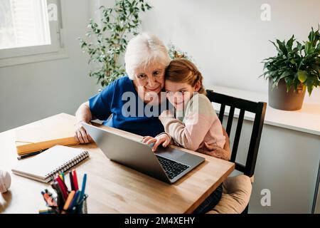 Lächelnde Großmutter hilft ihrer Enkelin beim Lernen mit einem Laptop auf dem Tisch zu Hause Stockfoto