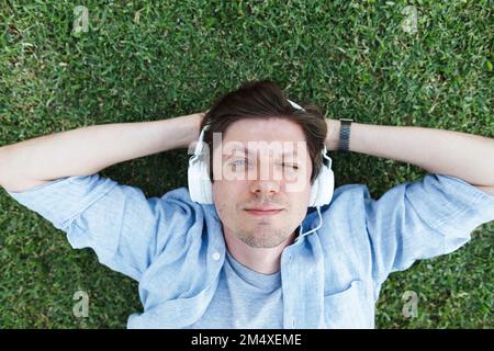 Lächelnder Mann, der Musik über Kopfhörer hört und sich im Gras entspannen kann Stockfoto