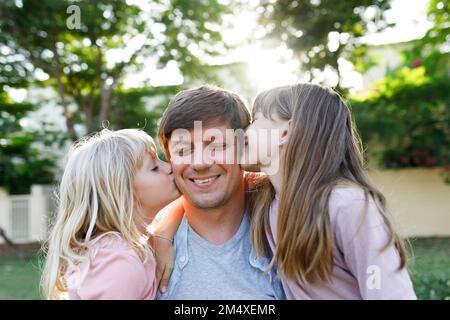 Lächelnder Vater mit Töchtern, die ihn im Park auf die Wange küssen Stockfoto