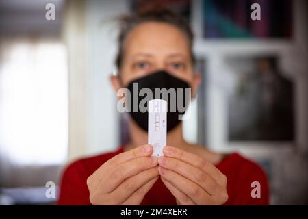 Frau trägt Gesichtsmaske, die ihren positiven Covid-19-Schnelltest zu Hause zeigt Stockfoto