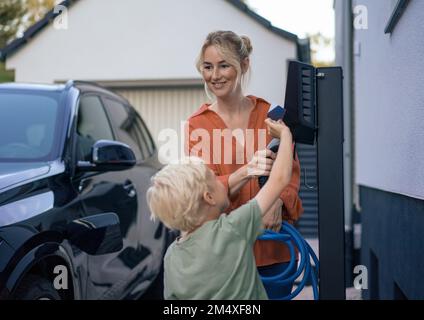 Ein Junge bezahlt per Kreditkarte von einer Frau, die den Stecker für das Ladegerät für Elektrofahrzeuge im Vorgarten hält Stockfoto