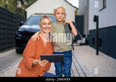 Mutter mit Tablet-PC, Sohn, der den Stecker des Ladegeräts für Elektrofahrzeuge im Vorgarten hält Stockfoto