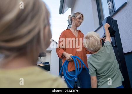 Ein Junge bezahlt per Kreditkarte von einer Mutter, die den Stecker für das Ladegerät für Elektrofahrzeuge im Vorgarten hält Stockfoto