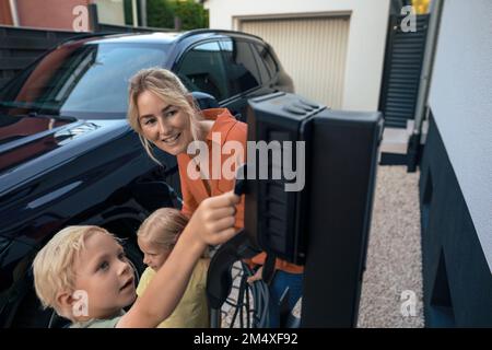 Sohn, der mit Kreditkarte bezahlt, von einer Mutter, die den Stecker für das Ladegerät für Elektrofahrzeuge im Vorgarten hält Stockfoto