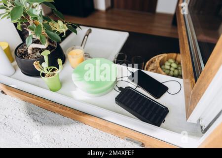 Mobiltelefon wird über eine Powerbank mit Solarbatterie auf der Fensterbank aufgeladen Stockfoto