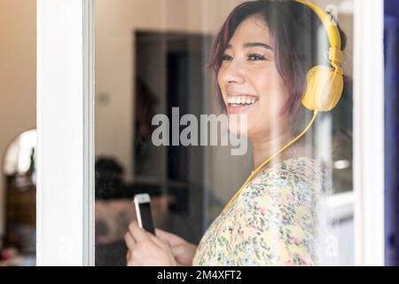 Glückliche junge Frau mit Kopfhörern, die durch ein Glasfenster gesehen wird Stockfoto