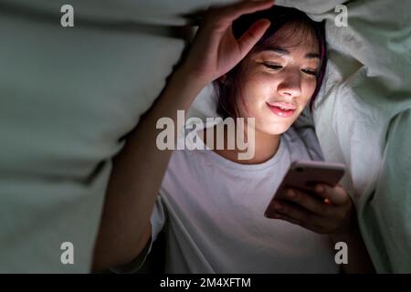 Lächelnde Frau, die zu Hause ein Smartphone unter der Decke benutzt Stockfoto