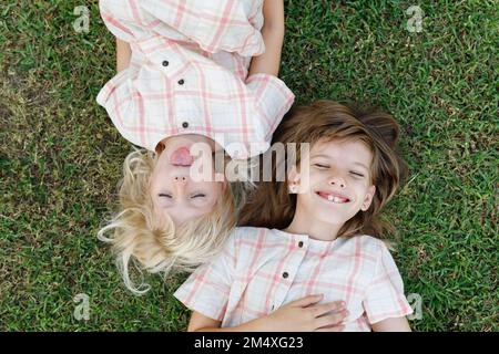 Süßes Mädchen, das die Zunge rausstreckt und von einer glücklichen Schwester auf dem Gras liegt Stockfoto