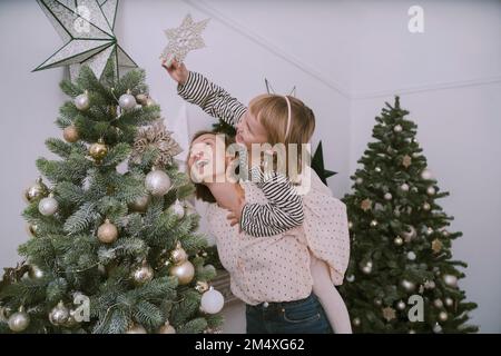 Tochter, die mit Mutter zu Hause einen Stern auf den Weihnachtsbaum legt Stockfoto