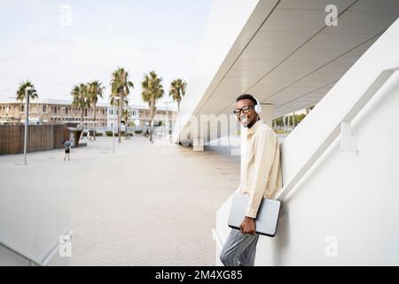 Lächelnder Mann mit Kopfhörern, der den Laptop an der Wand hält Stockfoto