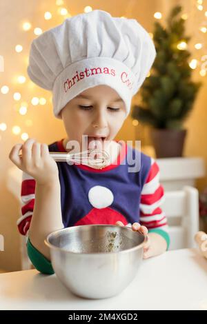 Ein Junge, der einen Kochhut trägt und in der Küche Teig auf einem Schneebesen leckt Stockfoto