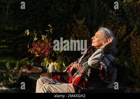 Seniorin sitzt im Herbstgarten in einem Sessel Stockfoto