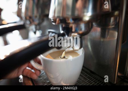 Die Hand des Baristas hält den Becher unter der Espressomaschine und gießt Milch ein Stockfoto