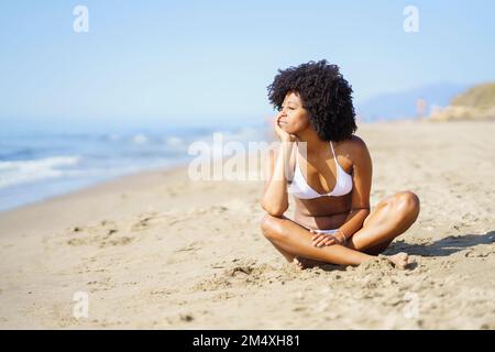 Rücksichtsvolle junge Frau, die am Strand auf Sand sitzt Stockfoto