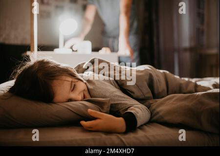 Ein glücklicher Junge schläft im Bett, während der Vater im Hintergrund das Licht ausschaltet Stockfoto