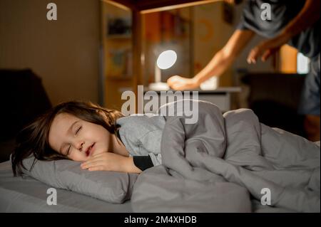 Der Sohn schläft im Bett, während der Vater das Licht im Hintergrund ausschaltet Stockfoto