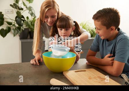 Lächelndes Mädchen hilft Schwester beim Sieben von Mehl durch Bruder zu Hause Stockfoto