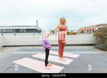 Glückliche Mutter und Tochter mit Händen an den Hüften beim Yoga auf dem Dach Stockfoto