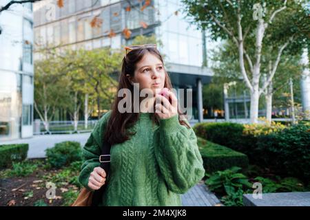 Rücksichtsvolle junge Frau, die Apfel vor dem Gebäude isst Stockfoto