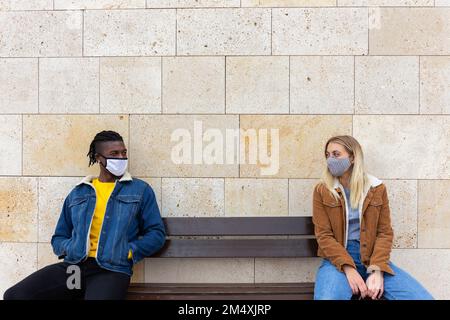 Ein junges Paar, das Gesichtsschutzmasken trägt und auf einer Bank sitzt Stockfoto
