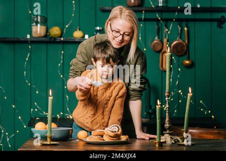 Lächelnde Mutter, die dem Sohn hilft, Puderzucker auf Zimtbrötchen in der Küche zu streuen Stockfoto