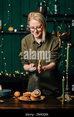 Lächelnde Frau, die Puderzucker auf Zimtbrötchen in der Küche streut Stockfoto