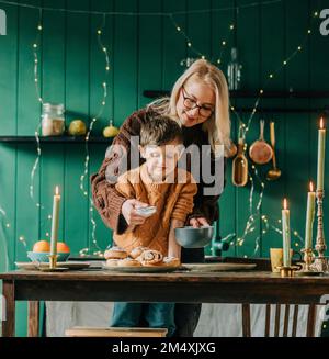 Mutter, die dem Sohn hilft, Puderzucker auf Zimtbrötchen in der Küche zu streuen Stockfoto