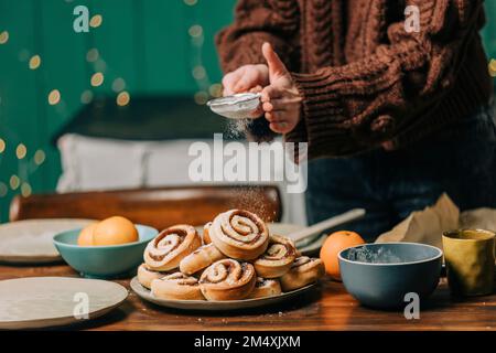 Hände einer Frau, die zu Hause Puderzucker auf Zimtbrötchen streut Stockfoto