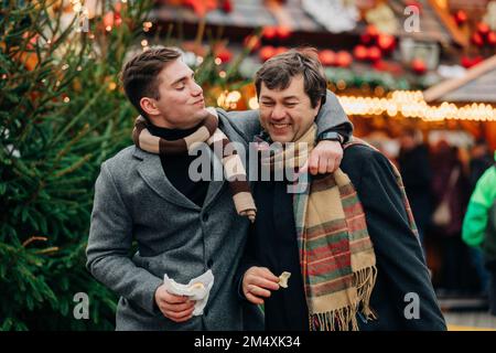 Lächelnder Mann, der Tdelnik isst, mit dem Arm um den Vater, genießt den Weihnachtsmarkt Stockfoto