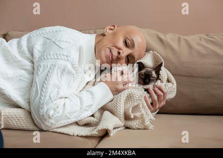 Kahlköpfige Frau, die die Sphynx-Katze umarmt, in eine Decke auf dem Sofa gewickelt Stockfoto