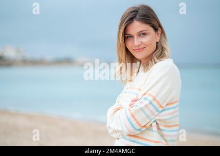 Lächelnde blonde junge Frau mit gekreuzten Armen, die am Strand stand Stockfoto