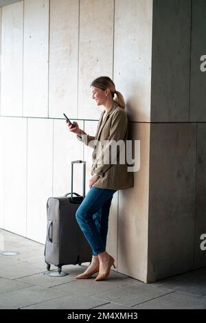 Lächelnde Geschäftsfrau, die ein Smartphone benutzt, wenn ihr Gepäck im Tunnel an die Wand lehnt Stockfoto