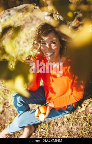 Glückliche Frau, die mit Orangenfrüchten unter dem Baum sitzt Stockfoto