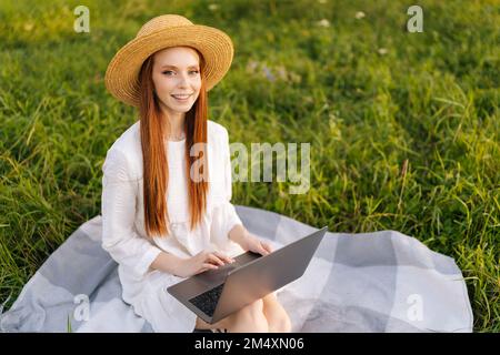 Porträt einer glücklichen, eleganten jungen rothaarigen Frau mit Strohhut und weißem Kleid, die auf Karo sitzt und mit einem Laptop in die Kamera schaut. Stockfoto