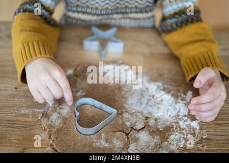 Hände eines Jungen, der Kekse mit herzförmigem Messer auf dem Tisch ausschneidet Stockfoto