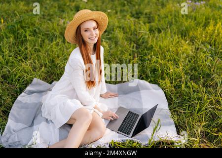 Blick aus der Vogelperspektive auf einen fröhlichen jungen rothaarigen Freiberufler mit Strohhut und weißem Kleid, der mit einem Laptop in die Kamera schaut. Stockfoto