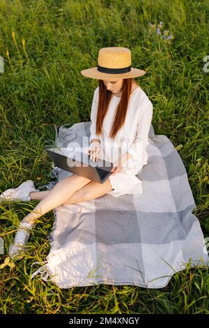 Vertikale Ansicht aus einem hohen Blickwinkel einer fokussierten Freiberuflerin mit Strohhut und weißem Kleid, die auf einem Laptop auf einem wunderschönen grünen Grasfeld sitzt. Stockfoto