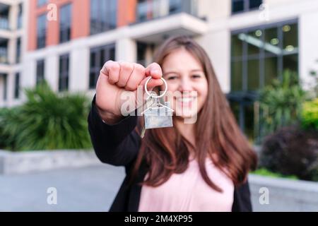 Glückliche junge Frau, die vor dem Gebäude die Hausschlüssel zeigt Stockfoto