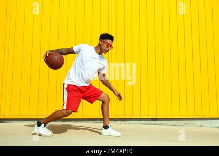Junger Mann, der mit Basketball auf dem Fußweg vor der gelben Wand spielt Stockfoto