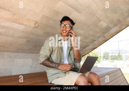 Lächelnder Freiberufler, der über ein Mobiltelefon spricht und mit einem Laptop auf der Bank sitzt Stockfoto