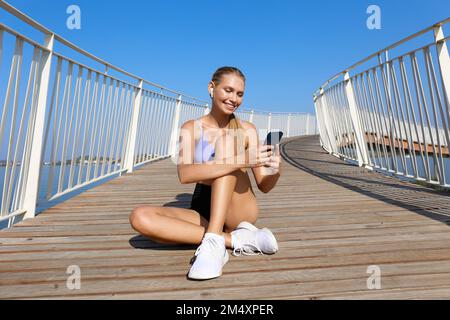 Glückliche Frau mit kabellosen in-Ear-Kopfhörern, die auf einem erhöhten Gehweg auf ein Mobiltelefon schaut Stockfoto